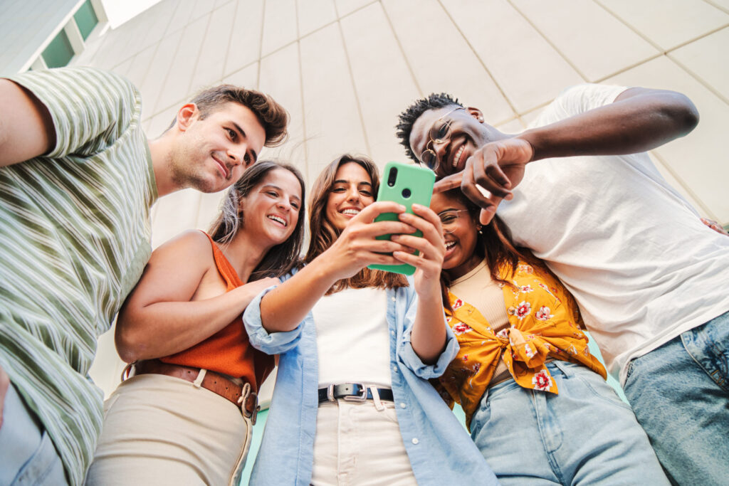 Low angle view of multiracial group of young friends enjoying and smiling using their mobile phone app at university campus. diverse teenagers having fun watching content on the social media outdoors
