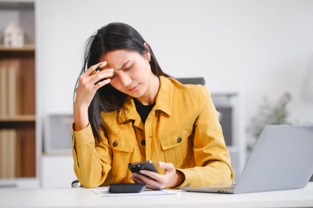A businesswoman works at her desk, looking at her phone and documents. she feels stressed and experiences a migraine due to workload, deadlines, and business pressure, affecting her productivity