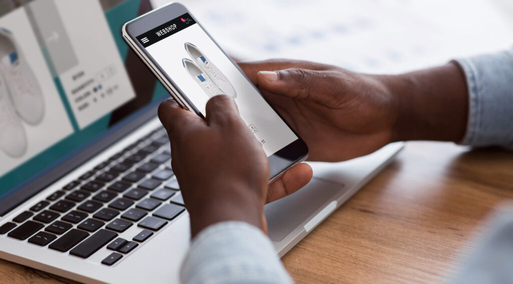 Black man shopping online with smartphone and laptop indoors, cropped